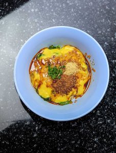 Overhead view of blue plastic bowl with noodles topped with yellow chickpea gravy, toasted peanuts, sesame seeds, herbs, and soy dressing