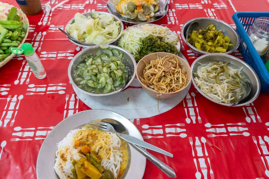 Table full of bowls with different vegetable based condiments inside (e.g. shredded banana blossom, pickled cucumber) and plates of thin rice noodles with curry on top.