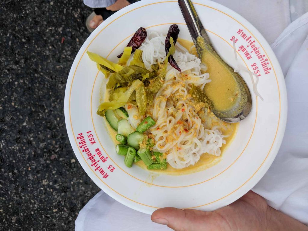 white plate with Thai text on the rim filled with white rice noodles, curry, and various toppings.