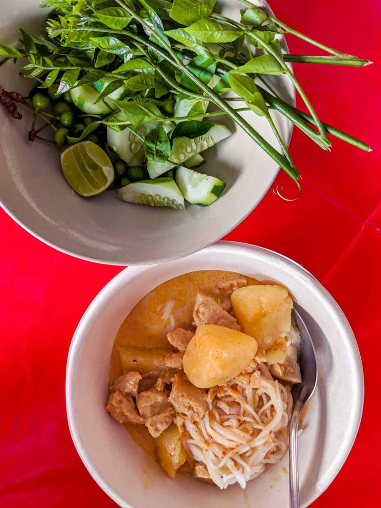 Bowl full of herbs and vegetables and another bowl with red curry noodles (including potatoes) on a red tablecloth